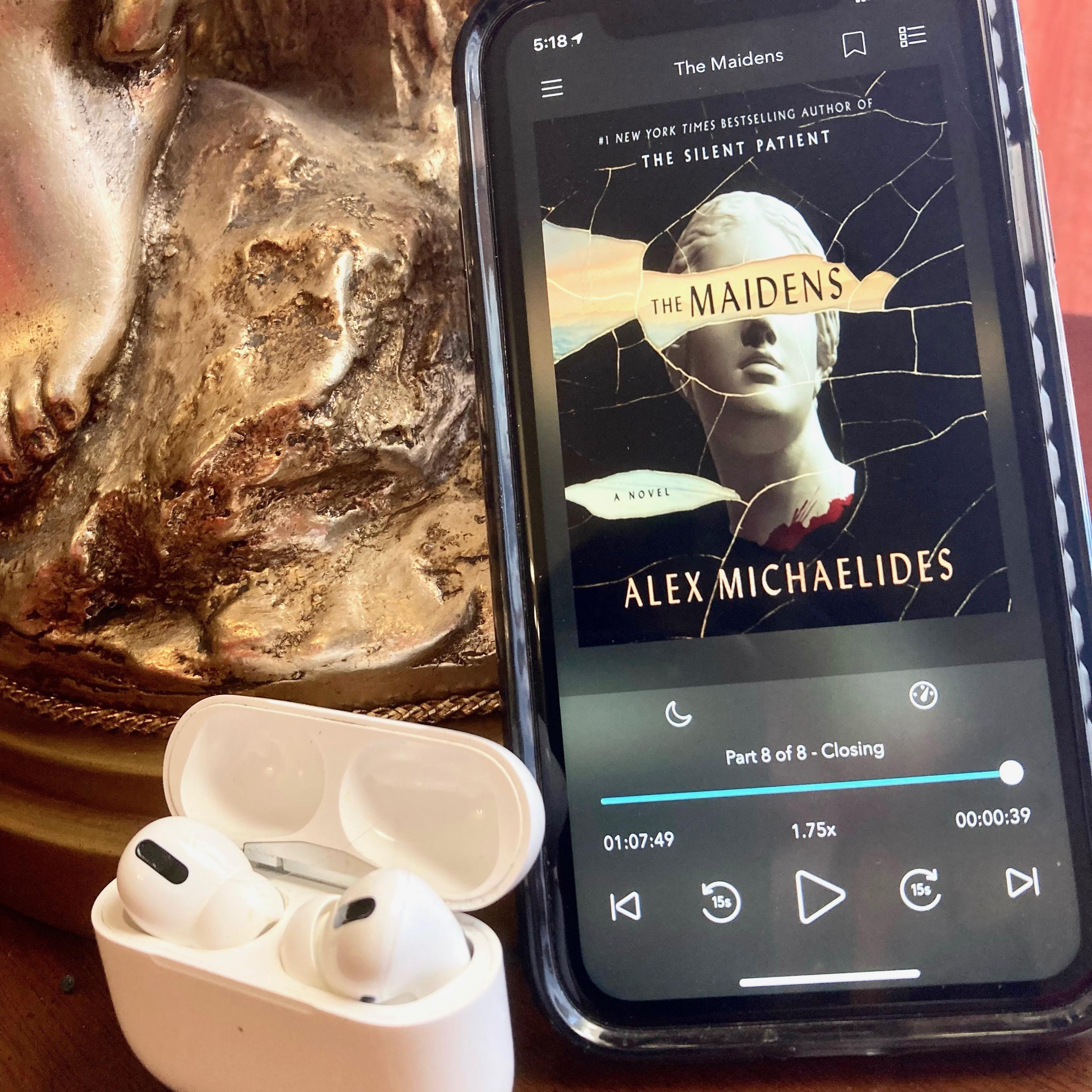 The Maidens by Alex Michaelides cover on iPhone next to AirPods and Lamp